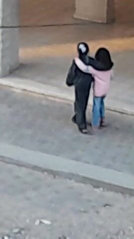 The video shows two people embracing in an outdoor setting. One person is wearing a black outfit and a mask, while the other is wearing a pink jacket and blue jeans. They stand close to each other, with the person in pink having their arms around the person in black. They are positioned near a white stone archway with a cobblestone ground. The lighting suggests it is daytime. There are other architectural elements and surfaces visible in the background, appearing slightly aged and textured.
