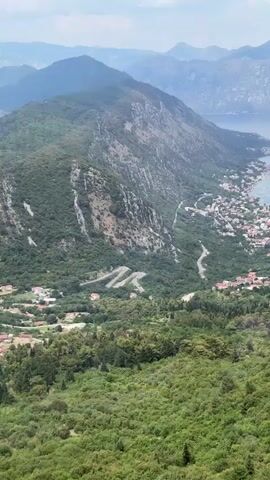 The video begins with a close-up of a modern, blue-accented cable car cabin, with the logo "XE energy" visible on its side. Inside, several people are seated, looking out at the scenery. The camera then pans to reveal a breathtaking panoramic view of a mountainous coastal landscape. Lush green vegetation covers the slopes, interspersed with winding roads and small clusters of houses. In the distance, the calm blue waters of a bay or sea stretch out, meeting the horizon line of more distant, hazy mountains. The overall impression is of a scenic cable car ride offering stunning views of a beautiful natural environment.