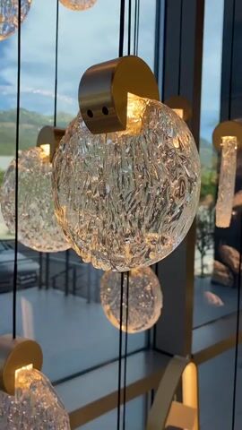 The video shows a close-up of several decorative glass pendant lights hanging in front of a large window. The lights have a textured, spherical glass design and are illuminated from within by a warm light source. They are suspended by thin black cords and attached to brass fixtures. Outside the window, a scenic view of mountains and sky is visible. The lighting suggests it is either early morning or late evening, with soft, diffused daylight. The camera is focused on the central pendant light, slowly panning around it, allowing the viewer to appreciate its intricate detail and the way light passes through the textured glass. Other similar pendant lights are visible in the background, creating a sense of a larger installation.