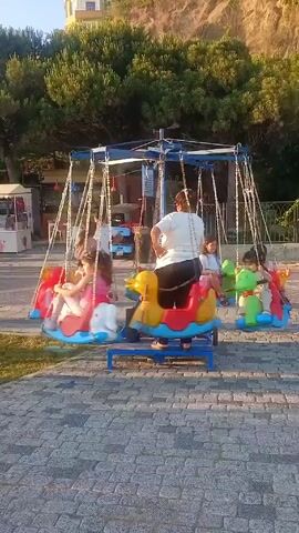 Here is a concise description of the video:

The video shows a children's ride in what appears to be a small park or outdoor area. Several young children are riding on a carousel-like contraption with seats shaped like colorful animals (duck, horse, and a dinosaur). The ride is in motion, swinging the children around. Two adults, one wearing a white t-shirt and the other one wearing a white shirt and red shorts, are nearby, appearing to supervise the children and the ride. The setting includes a brick-paved surface, a patch of grass, some trees, and a building in the background. An ice cream cart is also visible.