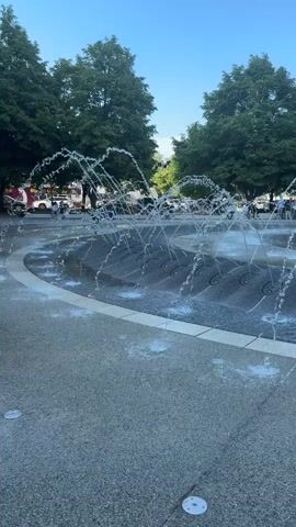 The video shows a public fountain in a park on a sunny day. The fountain is circular and has many small jets of water shooting upwards, creating an interactive water feature. The fountain's surface is dark gray and patterned. A concrete path surrounds the fountain. Several people, including children, are playing in and around the water, some running through the water jets. In the background, there are green trees, and a few buildings are visible in the distance. The sky is clear and blue. The camera is positioned at ground level, providing a wide shot of the scene.
