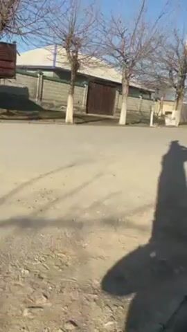 The video shows an old, light-blue tractor driving on an asphalt road. The tractor appears to be in poor condition, with missing panels and exposed engine parts.  There are several bare trees along the side of the road, along with a brick building with a fence and gate.  A blue car is also visible in the background. A long shadow of the person filming the video is visible, cast on the road.
