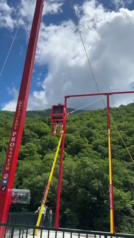 The video captures an outdoor amusement ride, specifically a giant swing, set against a backdrop of lush green mountains under a partly cloudy sky. The main structure consists of two tall red and yellow pillars supporting the swing mechanism. A person, likely a child, is seated in the swing, which is being pulled upwards by a rope. A man in a dark t-shirt and pants is visible on the platform, actively pulling the rope to raise the swing. Other spectators are seen on a viewing platform behind a white metal railing, watching the activity.

On the left red pillar, Cyrillic text is visible, reading "АБХАЗСКИЕ PROKACHELI" (Abkhazian swings/rockers), along with what appear to be social media logos (TikTok, Instagram). The scene takes place during the daytime, with clear visibility. The location is mountainous and heavily forested, suggesting a natural or scenic park setting, consistent with Bzypta, Abkhazia.