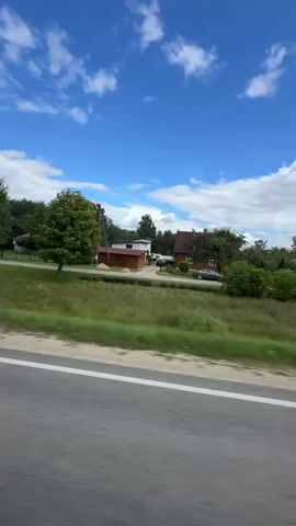The video is a time-lapse recording of a rural landscape taken from the perspective of a moving vehicle. The scene shows a road with a white line marking its edge. Beyond the road, there is a grassy area with trees and some buildings. The sky is blue with scattered white clouds. The camera is moving from left to right, suggesting the vehicle is traveling in that direction. There are no people visible in the video. The buildings appear to be houses and possibly some outbuildings. The overall impression is of a scenic drive through the countryside on a sunny day.