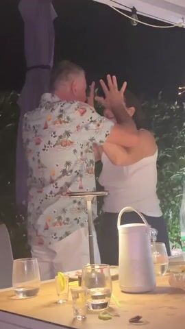 A man wearing a short-sleeved, light blue shirt with a tropical print and white pants stands facing a woman. The woman is wearing a white tank top and dark pants. She has her arms raised and appears to be touching the man's face. They are standing at a table outdoors at night. There are glasses and a speaker on the table.