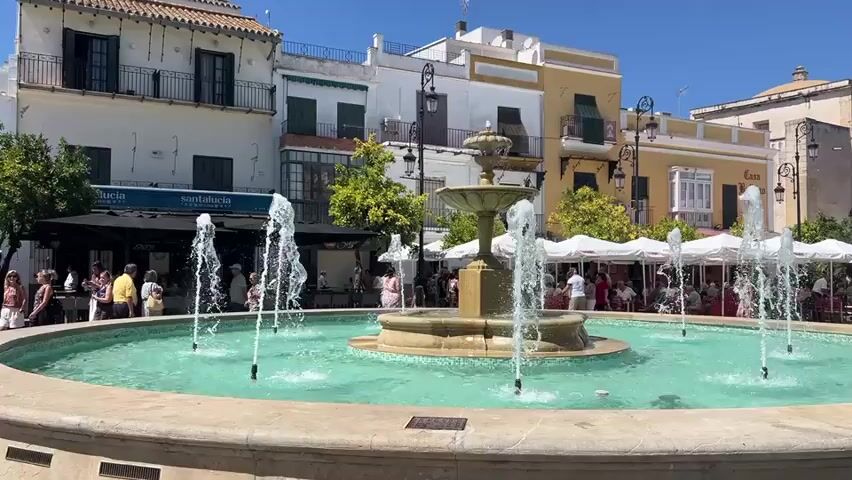 A wide shot captures a public square with a central fountain. The fountain features multiple water jets spraying into a circular basin filled with turquoise water. The basin is surrounded by a light-colored stone border. In the background, several buildings with white and yellow facades stand under a clear blue sky. Some buildings have balconies with dark railings. Trees with green foliage are visible between the buildings. In front of the buildings, cafes with white umbrellas and outdoor seating are set up. People are seated at the tables, and others are walking around the square. The ground of the square appears to be paved.