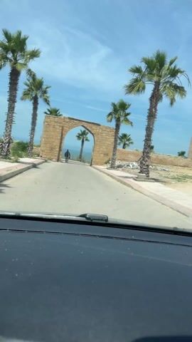 The video shows a car driving toward a stone archway on a sunny day. The road is paved, and palm trees line both sides. A man is walking through the archway away from the car. Beyond the archway, there is a glimpse of the ocean. There is also a ruined stone structure to the right of the road.