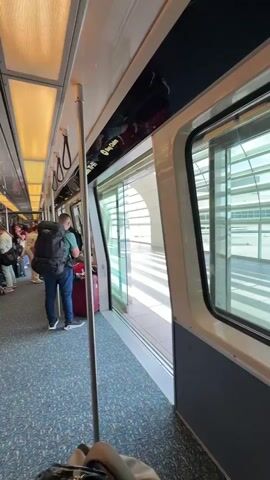 The camera looks out from the interior of an airport people mover train. People with luggage stand near the doors as the train is at a standstill.  The train has blue-speckled carpet, and beige lights cover the ceiling. Stainless steel poles run from the ceiling to the floor. The side windows show other people inside the airport. The door on the right has text that says “Sky Train.” The camera pans to the left while remaining still.