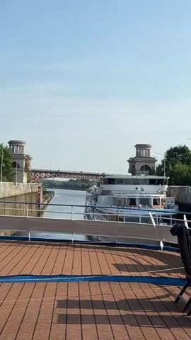 A camera pans across a river lock, showing a white cruise ship in the lock chamber. The ship has multiple decks with windows. In the background, a red bridge spans the river, and trees line the banks. The sky is clear and blue. The foreground shows part of the deck of a smaller vessel, with brown decking and blue trim.