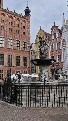 A video shows a fountain with a statue of Neptune in the center. The statue is of a muscular man holding a trident and standing on two horses. Water sprays from the fountain and from the mouths of four sculpted faces around the fountain's base. The fountain is surrounded by a black metal fence. Behind the fountain are several buildings with colorful facades, typical of European architecture. One building is tall and made of red brick, with arched windows. The sky is overcast.