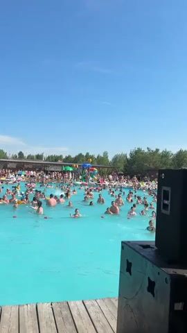 The video shows a crowded outdoor swimming pool on a sunny day. Many people are in the water, some on inflatable toys, and some are swimming. There is a wooden deck in the foreground with DJ equipment, including a laptop and a mixer. In the background, there are trees and buildings, suggesting a resort or water park setting. The sky is clear and blue.