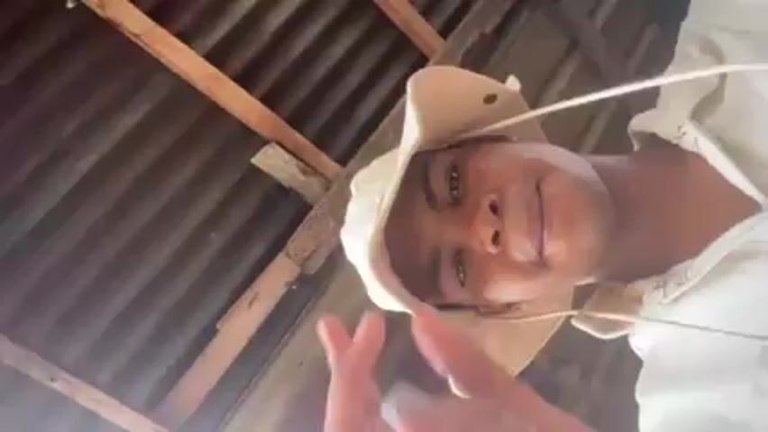 A low-angle video shows a Black man wearing a beige bucket hat and white shirt. He holds a rolled cigarette between his fingers and looks into the camera with a slight smile. Above him, there's a corrugated metal roof with wooden support beams. Sounds of a crowd and music are audible in the background.