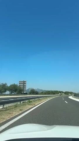 The video shows a view from inside a vehicle traveling on a highway on a clear, sunny day. The camera is pointed forward, capturing the road ahead, the roadside, and the sky. The highway is paved with asphalt and has white lane markings. A metal guardrail runs along the left side of the road. To the left of the highway, there is a grassy area with some bushes and trees. In the distance, there is a light-colored, multi-story building and a mountain range under a bright blue sky. The perspective suggests the vehicle is moving at a moderate speed.