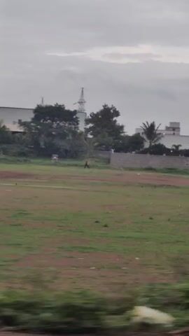 A group of young men are playing soccer on a grassy field. The sky is overcast, and the surrounding area appears to be industrial or semi-urban. There are several buildings and trees visible in the background. One man in a white shirt is running, while others are scattered across the field, some kicking a soccer ball.