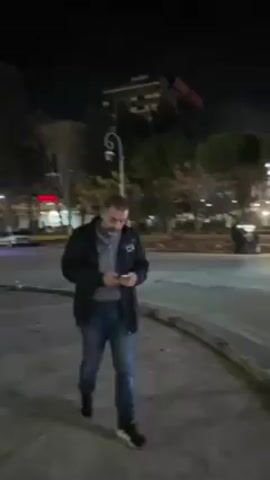 A man wearing a black jacket and jeans stands on a paved area at night, looking down at a small white object in his hands, possibly a phone or another electronic device. The scene is urban, with buildings, trees, and streetlights visible in the background. A road and a square are present with multiple vehicles and pedestrian carts with colored lights. The overall lighting is dim, but the area is illuminated by streetlights and the glow from the buildings.