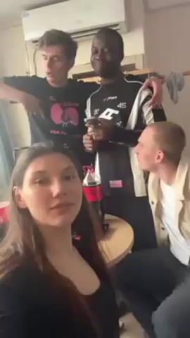The video shows four young adults, three males and one female, in what appears to be an indoor setting, possibly a dorm room. The female is in the foreground, facing the camera. Behind her, two of the males stand shoulder-to-shoulder. The male on the left wears a black T-shirt, and the male next to him, who is Black, wears a black-and-white racing-style jacket. The fourth male is seated and looking at the Black male. A small round table is present, holding a bottle of Coca-Cola. An air conditioning unit is visible on the wall above. The individuals are looking at the camera and smiling, while one of the males is looking at his phone. The lighting in the room is bright.