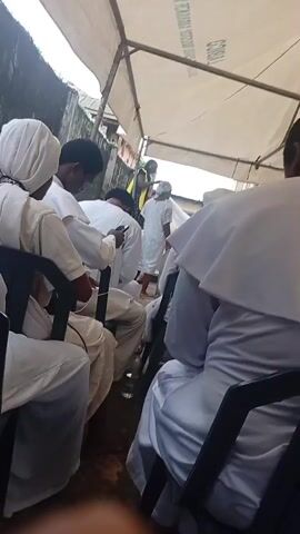 The video shows a group of people, mostly dressed in white, sitting on chairs under a makeshift tent. In the background, a woman in a yellow and black dress appears to be adjusting the headwear of a young person also in white. Some individuals in the foreground are looking at their phones. The environment appears to be outdoors, with a wall visible behind the tent. The overall atmosphere seems to be one of a gathering or event.