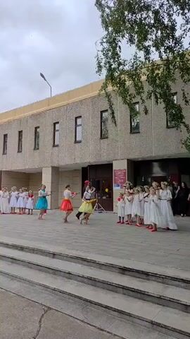 A group of young girls in white dresses and flower crowns stand in a line on a stone staircase in front of a building. Three girls in colorful tutus are dancing in front of them, one in blue, one in red, and one in yellow. They are performing a choreographed dance with synchronized movements. The building appears to be made of concrete with several windows. The sky is overcast.