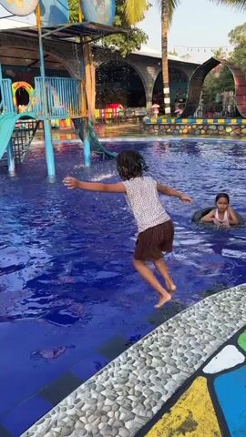 The media file depicts a sunny day at a water park in Kebonsari, Indonesia. The scene centers on a bright blue swimming pool with children enjoying themselves.

Two young girls are the primary subjects. One girl, in the foreground, is captured mid-splash as she jumps into the water, her arms outstretched and her body submerged. She is wearing a patterned swimsuit. Another girl is visible further back in the pool, sitting inside a black inner tube.

The pool area features a vibrant blue tiled edge with a textured, stone-like border. In the background, a multi-colored water slide structure with blue accents stands prominently. The slide has a green flume and a yellow platform. Other elements of the water park are hinted at, including what appears to be covered seating areas and arched structures in the distance. Palm trees are visible, suggesting a tropical climate.

The time of day appears to be daytime, likely morning or early afternoon, given the bright sunlight casting shadows and illuminating the water. The weather seems pleasant and warm, suitable for swimming. The overall atmosphere conveyed is one of fun and joyful activity, with the children's energetic play suggesting happiness and excitement.

No visible text is present within the frame.