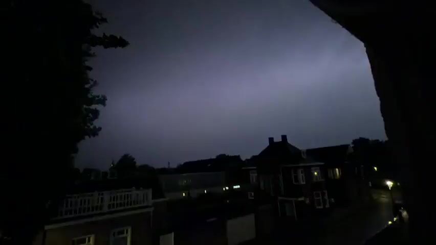 The video captures a dark, nighttime scene with a stormy sky. A faint, purplish glow emanates from the sky, indicating distant lightning. Below the sky, the silhouettes of houses and trees are visible. The overall atmosphere is dark and somewhat ominous, suggesting a severe weather event.