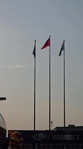 The video shows an outdoor event with people gathered. In the foreground, a group of women in light-colored dresses are holding a long red ribbon. To the left, there is a traditional Chinese lion dance costume with a performer visible. Behind the women, a large banner with the word "CARRY" and Chinese characters is displayed on a building. Three flags are visible in the background, one of which is the Chinese flag. The event appears to be a ceremony or celebration, possibly a product launch or opening. The lighting suggests it is either early morning or late afternoon.