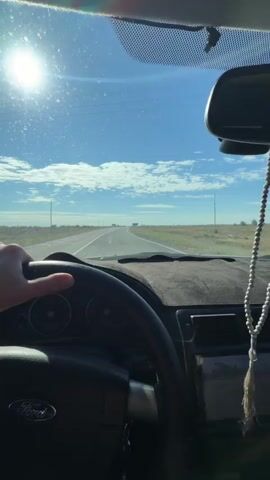 The video shows a first-person perspective from inside a car driving on a highway. The driver's left hand is visible on the steering wheel, which has a Ford logo on it. The dashboard is covered with a dark fuzzy material. The road ahead is straight with white lane markings. The sun is shining brightly in the upper left corner of the frame, creating lens flare. The sky is blue with scattered white clouds. In the distance, there are utility poles and signs along the side of the road. A beaded string hangs from the rearview mirror. The overall scene depicts a sunny day drive on an open road.