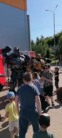 A full shot shows Optimus Prime and Bumblebee mascots in a large crowd of children and adults. Optimus Prime is on the left and has a red chest, black torso, and blue metallic pants. He is wearing a black helmet. Bumblebee is on the right and is yellow and black. The crowd is gathered around both mascots, and a man is standing in the middle of the crowd with a microphone. The event appears to be outdoors.