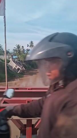The video shows a river with muddy water under a partly cloudy sky. On the left bank, a house has partially collapsed and is leaning precariously into the river. The riverbanks are lined with palm trees and other vegetation. In the foreground, a red metal bridge structure is visible, and on the bridge, a flag on a bamboo pole is seen. In one frame, a person on a motorcycle with a helmet is briefly visible riding on the bridge. The overall scene suggests a rural or semi-rural area affected by natural events, possibly flooding or erosion, that has damaged the house.