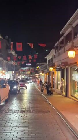 The video captures a nighttime street scene. Red flags with black eagles are strung across the cobblestone street, suggesting a national celebration or event. Cars and motorcycles navigate the street, with their headlights illuminating the path. Shops line the right side of the street, their lights casting a warm glow on the pavement. A woman in a floral dress is visible in the foreground, holding up a smartphone to record the scene. People are seen walking along the sidewalk, some with strollers, and others standing outside the shops. The overall atmosphere is lively and bustling.