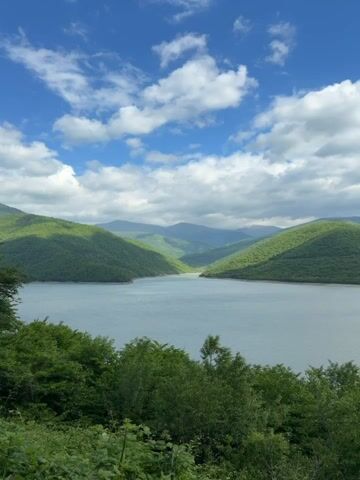 The video shows a wide shot of a lake surrounded by green, forested hills under a blue sky with scattered white clouds. The lake's water is a calm grey-blue. The hills are covered in lush greenery. In the foreground, there are trees and shrubbery, providing a sense of depth. The sky is bright with white, fluffy clouds and patches of blue. There is no visible human activity in the frame.
