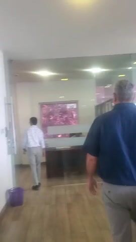 The video opens showing a man in a plaid shirt and light-colored pants walking away from the camera into an office space. He is holding something in his right hand. A purple trash can is visible to the left. The camera then pans to show a man in a blue shirt and light-colored pants walking away from the camera. He is also holding something in his right hand. The video then shows a glimpse of a ceiling light and then a staircase with a metal handrail. The final shot is a selfie of a woman wearing a patterned headwrap and a yellow and brown patterned shirt, making a kissing face.