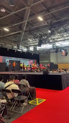 A group of people are performing on a stage with drums in front of an audience. The stage is set up indoors, likely in an exhibition hall, as indicated by the high ceiling with exposed industrial beams, lighting rigs, and large speakers. The backdrop of the stage features colorful graphics with the word "SAKURA" repeated multiple times, suggesting a theme related to Japanese culture or a specific event. To the right of the stage, a green sign reads "SORTIE DE SECOURS," which translates to "EMERGENCY EXIT" in French, a common sight in public venues in France.

The performers, a mix of adults and possibly older children, are dressed in vibrant clothing. Several individuals are actively playing large, red taiko drums, holding drumsticks and demonstrating energetic movements. Other individuals on the stage appear to be directing or participating in the performance. The audience is seated in chairs, facing the stage, with some individuals visible in the foreground, also seated. The overall atmosphere appears to be that of a lively cultural performance or festival. The lighting suggests it is daytime, but the indoor setting makes it difficult to determine external weather conditions. The presence of the "EMERGENCY EXIT" sign confirms the location is within France, specifically Villepinte, as per the provided context.