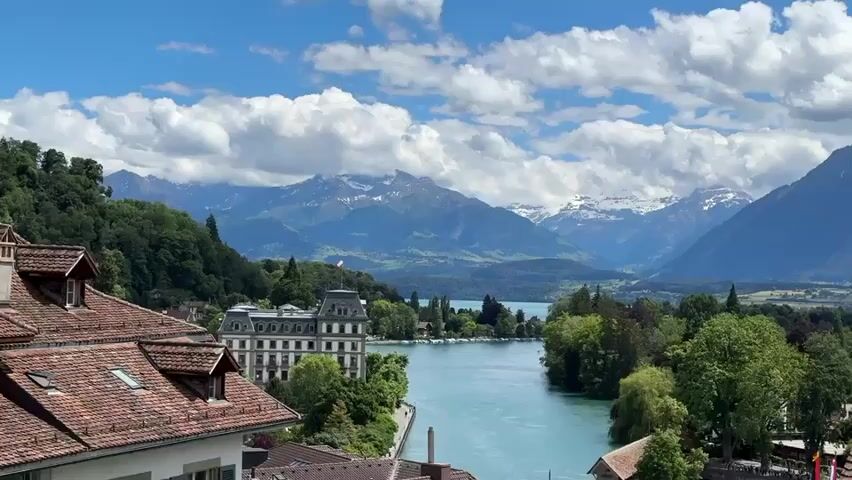 A panoramic video captures a scenic view of a turquoise lake with snow-capped mountains in the background under a partly cloudy sky. In the foreground, the red tile roofs of buildings are visible, with a large white building situated on the left bank of the lake. Trees line the lake's edge, and a small town can be seen in the distance. The camera pans slowly from left to right, offering a continuous vista of the natural landscape and the urban environment. No people or specific actions are discernible.