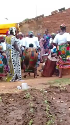 The video shows a gathering of approximately 20 women in an outdoor space. Most of the women are wearing colorful, patterned dresses and headwraps. They appear to be engaged in some form of celebration or dance. Some are standing and moving to the music, while others are seated. A few women are playing a large, wooden drum, providing the rhythm for the event. There’s a yellow canopy overhead. The ground is a mix of dirt and grass, indicating a natural or rural setting. In the foreground, tilled dirt can be seen. A brick building or wall is visible in the background. There is a white plastic cup on the ground. The overall atmosphere appears lively and communal.