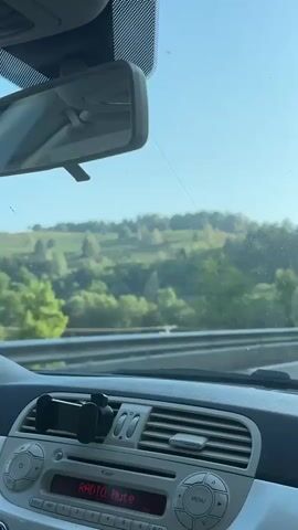 The video shows the interior of a car driving on a road. The dashboard is visible in the foreground with a phone mount attached. The windshield shows a bright, clear sky with trees and rolling hills in the distance. A rearview mirror is also visible, reflecting the interior of the car. A lamppost and a signpost are seen outside the car. Another car is visible ahead on the road. The scene appears to be a rural or suburban setting.