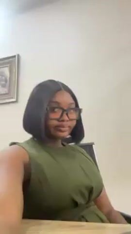 A woman with dark skin, wearing black-framed glasses, a sleeveless green dress, and a short, dark bob hairstyle, sits in a chair at a desk. A framed picture hangs on the cream-colored wall behind her. The camera appears to be handheld and slightly below eye level. The woman looks directly at the camera, then briefly glances to her right.