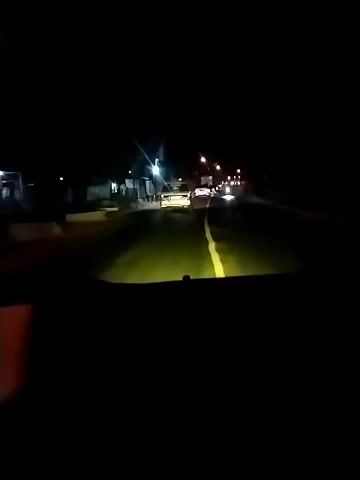 A first-person perspective video recorded at night shows a vehicle traveling on a road. The headlights of the vehicle illuminate the road ahead, creating a bright yellow glow. Several other vehicles are visible on the road, some with their headlights on, and others with taillights illuminated. There are streetlights and lights from buildings visible along the side of the road. The road appears to be paved and has a yellow line marking the center. The video captures the movement of the vehicle and the surrounding environment in low-light conditions.