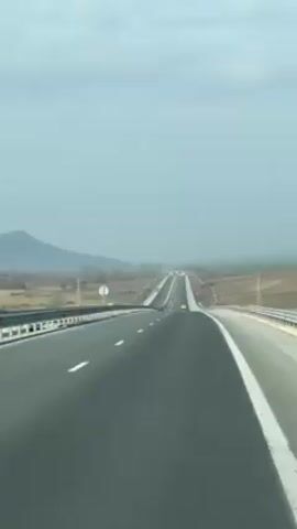 The video shows a car traveling down a highway on a cloudy day. There is a mountain in the distance. The landscape on either side of the highway consists of fields. A yellow car is far ahead.