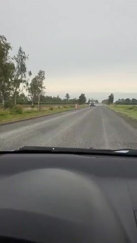 The video shows a view from inside a vehicle looking out at a road. A white truck with "MAN" written on the front is driving towards the camera. The road is wet and appears to have a textured surface. The sky is overcast. There are trees and fields on either side of the road. Several traffic markers are visible along the side of the road. The truck is the primary focus of the initial part of the video, and then the view shifts to a car driving away from the camera on the same road.