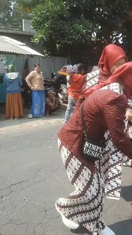 The video shows a group of people, predominantly women and children, walking in a procession or parade. They are all dressed in traditional Indonesian attire, consisting of red batik shirts and pants or skirts, and red headscarves (hijabs). Some of the children are holding small Indonesian flags. The procession appears to be taking place outdoors on a paved road, with trees and buildings visible in the background. The lighting suggests it is daytime, possibly sunny. The individuals are moving in the same direction, with some adults appearing to supervise or escort the children. The overall impression is of a cultural event or celebration.