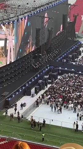 A high-angle, long shot captures a large crowd of people gathered in front of a stage within a stadium. The stage features two large screens displaying images, and a choir is visible on stage. The stadium seating, filled with more spectators, forms a curved backdrop. Several security personnel in black and yellow vests are scattered among the crowd. A large speaker hangs from a metal structure on the right side of the stage. A pathway separates the main crowd from the stage area.