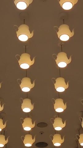 The video shows a ceiling fixture composed of rows and columns of white teapot-shaped lights. Each teapot hangs from a thin wire, and the light shines from the bottom of the pot. Between the rows of lights are round, dark gray vents and a smoke detector. The lighting appears warm.