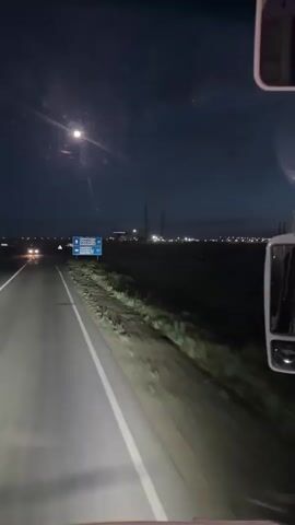 The video shows a view from inside a vehicle at night. A dark road stretches ahead, illuminated by headlights and a faint glow from streetlights or distant buildings. The sky is dark with a visible moon. On the right side of the road, there is a blue signpost with white text and arrows, indicating directions. The vehicle appears to be in motion, moving along the road. The landscape on either side of the road is dark and appears to be open land, possibly fields or undeveloped terrain. In the distance, there are faint lights and structures, suggesting a populated area or industrial site. The overall atmosphere is one of traveling at night.