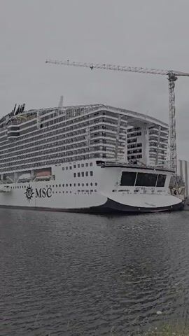 This media file captures a large cruise ship, partially under construction, docked in a body of water. The ship is predominantly white with a dark blue waterline. The words "MSC" are prominently displayed on the side of the hull, along with a circular logo. The upper decks of the ship are visible, showing rows of windows and what appears to be ongoing construction work, indicated by scaffolding and exposed structural elements. A large crane stands in the background, further suggesting a shipyard or construction site. The sky is overcast, implying a cloudy or gray day. The water is calm, with some ripples visible. The overall scene suggests a location in a port or shipyard, likely in Saint-Nazaire, France, given the context. There are no people clearly visible in the frame, but the presence of the ship implies a considerable amount of human activity associated with its construction and operation.