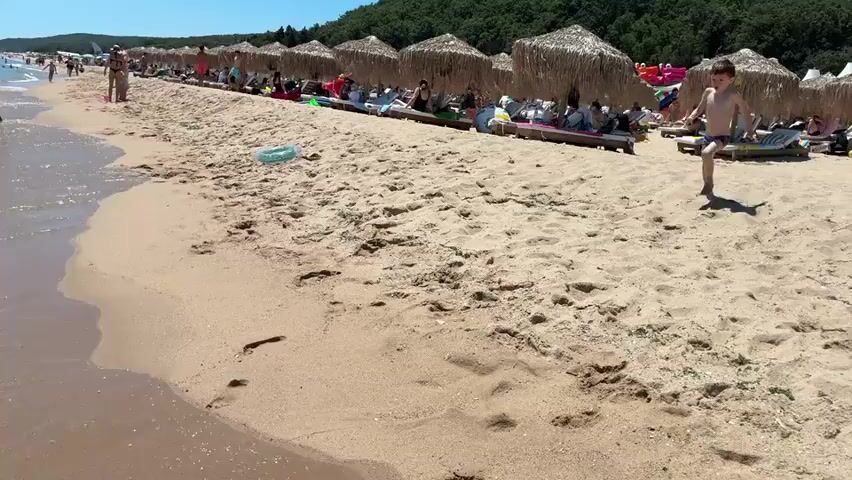 A boy runs into the ocean on a crowded beach. People are lying on lounge chairs under thatched umbrellas. The water is clear and the sky is blue. The boy splashes into the water, then gets out and runs back onto the beach.