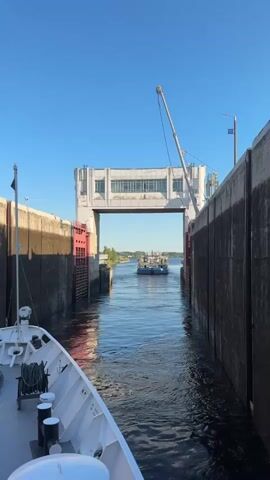 A video shows a blue boat with white structures on its deck, including what appear to be cabins and containers, moving through a lock. The boat is traveling away from the camera, towards a large, white, rectangular building with many windows. The lock is flanked by two tall, dark grey concrete walls. On the left side of the lock, a portion of a white boat is visible in the foreground. The sky is clear and blue.