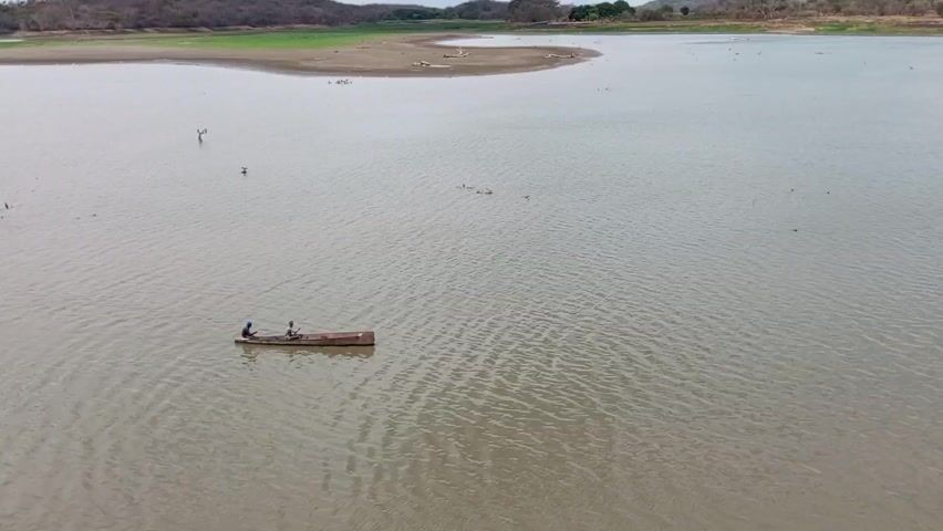 The video presents a wide, steady aerial shot of a landscape dominated by a large body of water. Two individuals are visible in a small, rustic boat, positioned centrally in the frame and moving slowly across the water's surface. The water is murky and still, reflecting the overcast sky. In the background, a partially submerged land mass, likely a peninsula or an island, exhibits a barren, dried-out appearance with scattered vegetation and debris. Further beyond, the shoreline is lined with low-lying, forested hills, adding depth to the scenery.  The overall setting suggests a remote or rural area, possibly experiencing drought or low water levels.