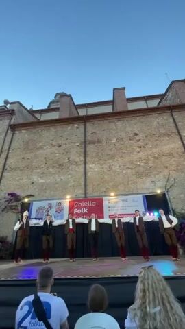 The video shows a group of men performing a folk dance on a stage in front of a stone building. They are all wearing similar traditional clothing: brown pants, white shirts, vests, and red sashes. The stage is set up outdoors, with a banner in the background that reads "Calella Folk Festival." The sky is clear and blue, suggesting it is daytime or early evening. There are lights illuminating the stage and some foliage on the left side. In the foreground, the backs of several audience members are visible, indicating they are watching the performance. The camera is positioned from behind the audience, looking towards the stage. The dancers are in sync, with their arms extended to the sides and one leg bent at the knee. The overall atmosphere appears festive and cultural.