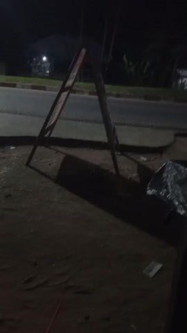 The video, set at night in what appears to be Mbribit, Nigeria, shows a dimly lit street scene. The primary focus is an A-frame sign standing on a dirt ground, casting a long shadow to the left. The sign's surface is not clearly legible.

In the background, a paved road is visible, with a red curb line. Vehicles occasionally pass by, indicated by blurred lights. A person wearing shorts and a dark shirt walks from right to left behind the A-frame sign and then out of frame. Moments later, another person, largely obscured by the right edge of the frame, also walks away.

On the ground near the sign, there is a silver, foil-wrapped object and some discarded paper or plastic. Beyond the road, there are dark silhouettes of buildings or structures and some sparse vegetation. Artificial light sources, likely streetlights, are visible in the distance, illuminating parts of the scene and creating reflections on the wet road surface. The overall atmosphere is dark and quiet.