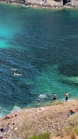 A high-angle, eye-level shot shows a body of water with several people swimming and sunbathing. The water is a clear, vibrant turquoise transitioning to deep blue. Rocky cliffs surround the water, with a small, pebbly beach on the right side of the frame. Several individuals are in the water, some swimming, others floating. On the beach, a few people are lying on towels, sunbathing. To the left, a group of people are gathered on the rocks, with one person standing on a paddleboard in the water. A pine tree with green foliage partially obscures the view on the left side of the frame. The sun is shining brightly, casting shadows and illuminating the scene.
